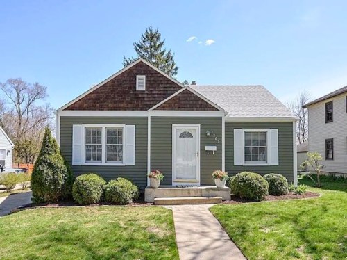 A charming green house with white shutters, a front porch, and landscaped yard on a sunny day.