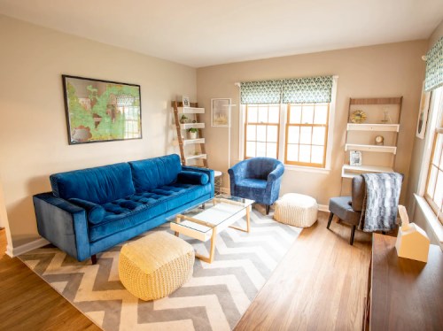 Cozy living room featuring a blue sofa, armchair, coffee table, and decorative shelves with natural light from windows.