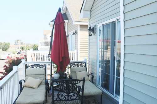 A cozy balcony with two lounge chairs, a table, and a red umbrella, surrounded by light-colored siding.