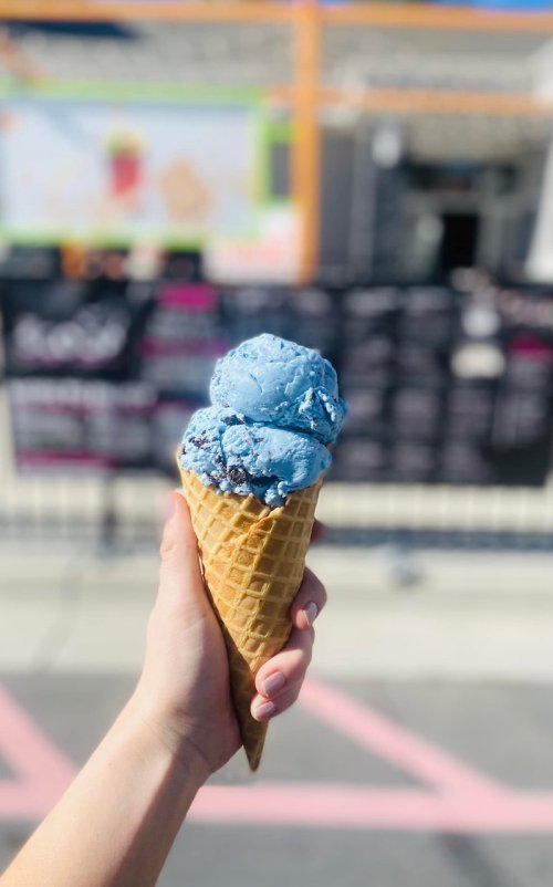 A hand holding a cone with two scoops of blue ice cream against a blurred background of a shop.