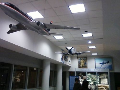 Two model airplanes hang from the ceiling in an aviation history exhibit, with visitors observing the displays below.