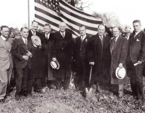 A group of men in suits and hats stands in front of an American flag, with a shovel in the foreground.