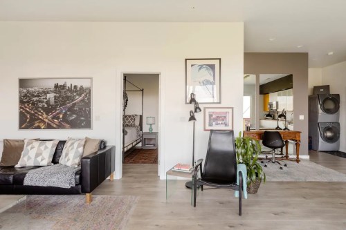 Modern living space featuring a black sofa, artwork, and a workspace with a desk and laundry area in the background.