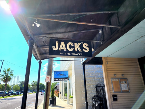 Sign for "Jack's by the Tracks" under a covered walkway, with palm trees and a street visible in the background.