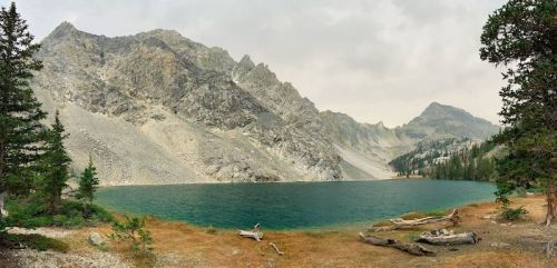 A serene lake surrounded by rocky mountains and trees, with a hazy sky above.