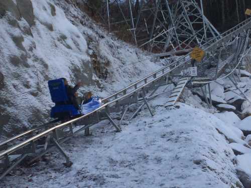 A person rides a sled down a snowy track beside a steep slope and metal structures.