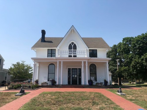 A large white Victorian-style house with a front porch, surrounded by green grass and trees under a clear blue sky.