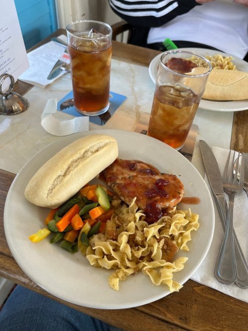 A plate with chicken, noodles, mixed vegetables, and a roll, accompanied by two glasses of iced tea on a table.