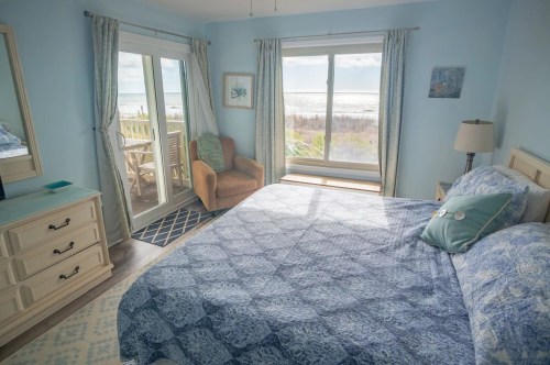 Cozy bedroom with a blue theme, featuring a large window overlooking the beach and a comfortable chair by the window.
