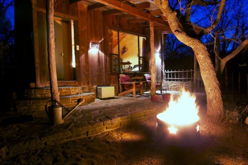 A cozy cabin at night with a glowing fire pit in front, surrounded by trees and a peaceful outdoor setting.