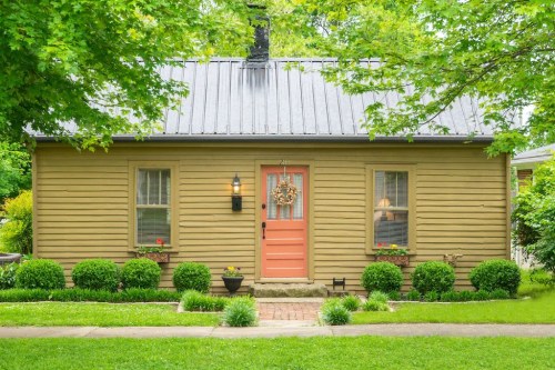 A charming yellow house with a pink door, surrounded by green bushes and a well-maintained lawn.