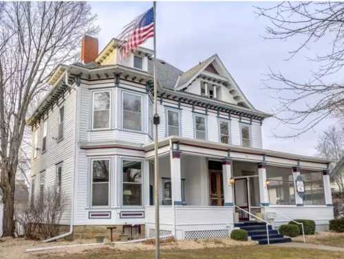 A large, historic white house with a porch, featuring an American flag and surrounded by bare trees.