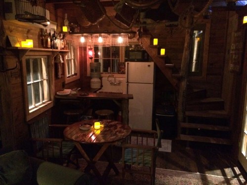 Cozy cabin interior with wooden decor, a small dining table, a fridge, and warm candlelight ambiance.