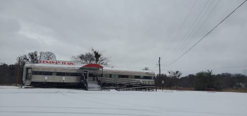 A vintage diner sits in a snowy landscape under a cloudy sky, with a clock sign and empty outdoor seating.