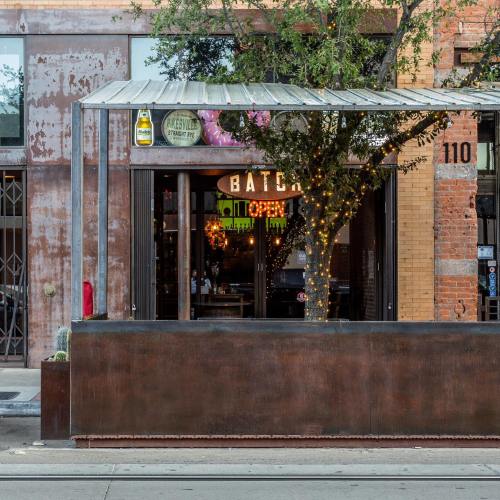 A rustic bar named "Batch" with an open sign, surrounded by greenery and string lights, in an urban setting.