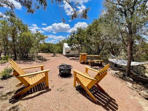 A serene outdoor space with two yellow chairs, a fire pit, a hammock, and lush greenery under a blue sky.