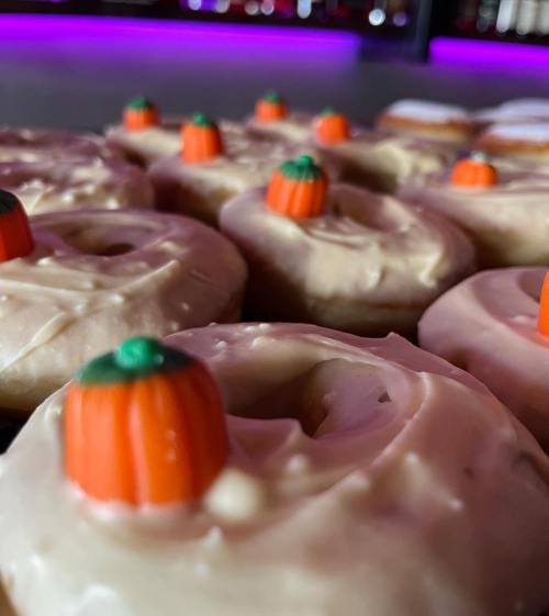 Close-up of frosted donuts topped with small orange pumpkin candies, set against a purple-lit background.