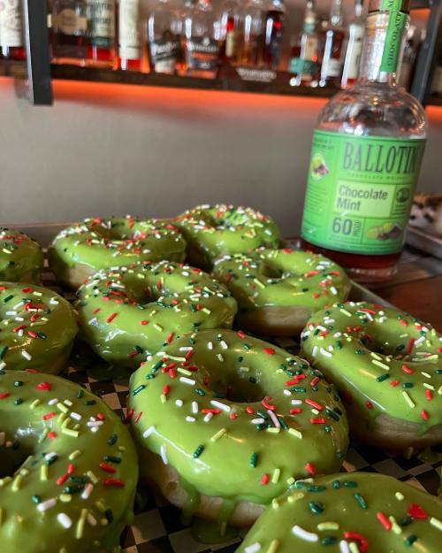 A tray of green frosted donuts topped with sprinkles, next to a bottle of Ballotin Chocolate Mint whiskey.