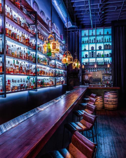 A stylish bar with illuminated shelves of liquor, wooden counter, and cozy seating in a dimly lit atmosphere.