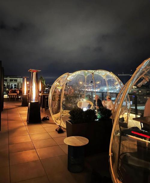 Rooftop setting at night with transparent bubble seating, heaters, and city views under a cloudy sky.