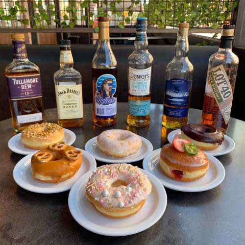 A variety of donuts on plates with several whiskey bottles arranged behind them on a table.