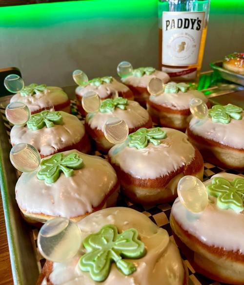 A tray of festive donuts topped with green icing and shamrock decorations, alongside a bottle of Paddy's whiskey.