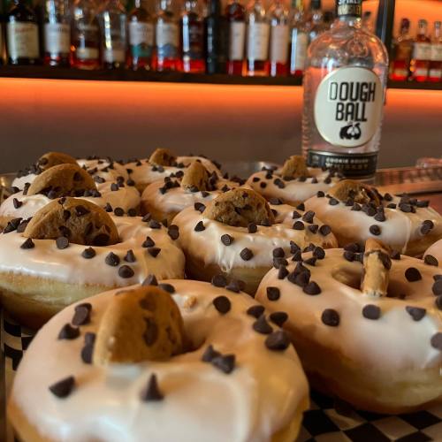 A close-up of donuts topped with icing, chocolate chips, and cookie pieces, with a bottle of Dough Ball in the background.