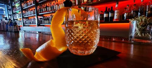 A crystal glass of cocktail garnished with a twist of orange peel, set on a bar with a colorful backlit shelf of bottles.