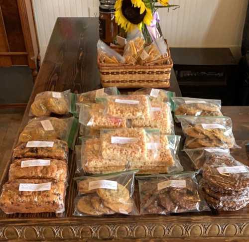 A wooden table displays various packaged baked goods, including cookies and bars, with a sunflower in the background.