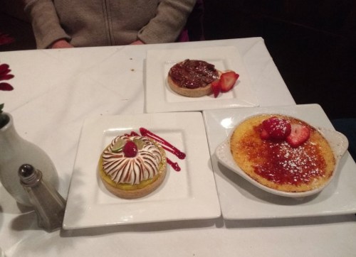 Three desserts on white plates: a fruit tart, a chocolate tart, and a crème brûlée, garnished with strawberries.