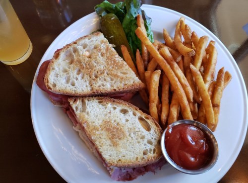 A plate with a toasted sandwich, pickles, lettuce, and a side of crispy fries with a small cup of ketchup.