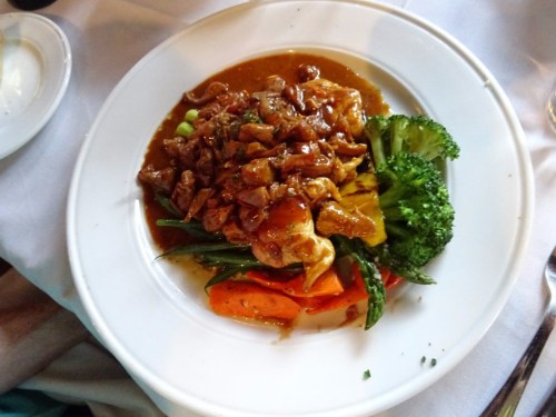 A plate of chicken with a savory sauce, served with broccoli, green beans, and carrots on a white tablecloth.