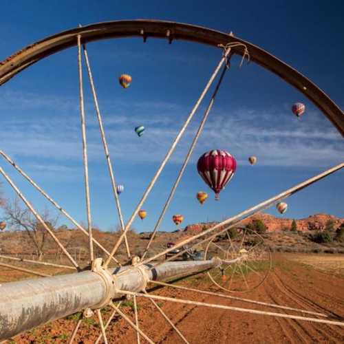 Colorful hot air balloons float above a field, framed by a large metal wheel against a clear blue sky.