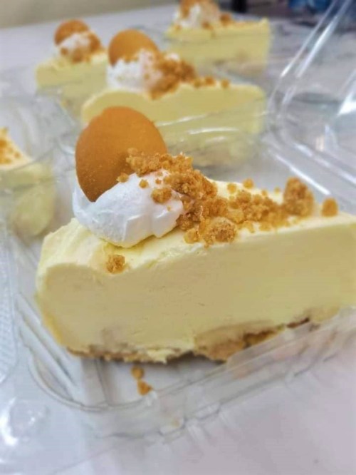 Slices of creamy yellow dessert topped with whipped cream, crushed cookies, and a mini cookie on a clear container.