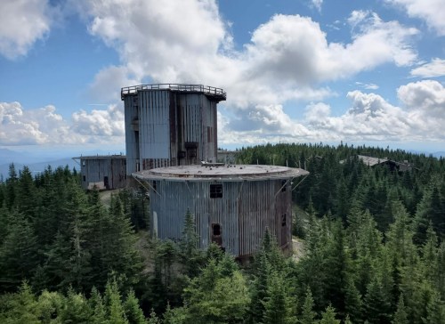 Two large, rusted structures surrounded by dense green forest under a partly cloudy sky.