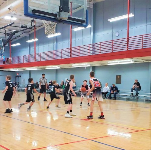 A youth basketball game in a gym, with players in black and white jerseys competing for the ball. Spectators watch from the sidelines.