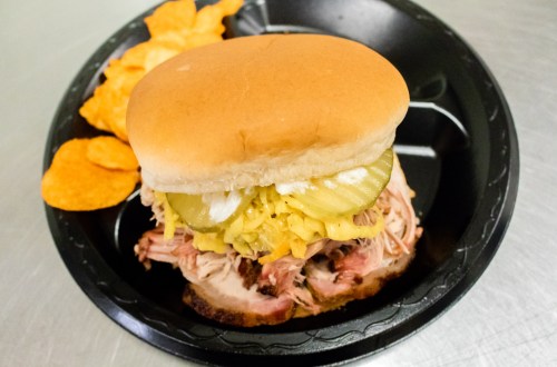 A pulled pork sandwich with pickles and mustard, served on a bun with a side of potato chips on a black plate.