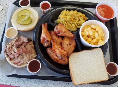 A tray with BBQ chicken wings, pulled pork sandwich, macaroni and cheese, pickles, and two slices of bread.