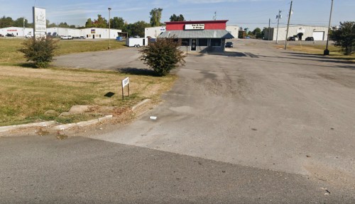 A vacant lot with a paved road leading to a building with a red roof and surrounding grassy areas.