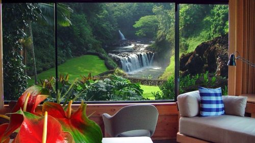 A serene view of a waterfall and lush greenery through a large window, with a cozy seating area in the foreground.