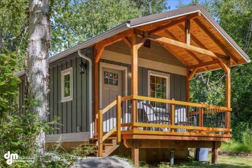 A small, modern cabin with a wooden porch, surrounded by trees and greenery.