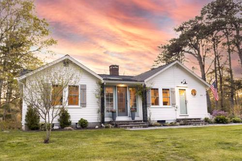 A charming white house with a porch, surrounded by greenery, under a vibrant sunset sky.