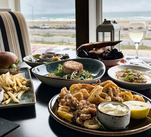 A table with various dishes, including seafood, a burger, fries, and a glass of wine, with a beach view in the background.