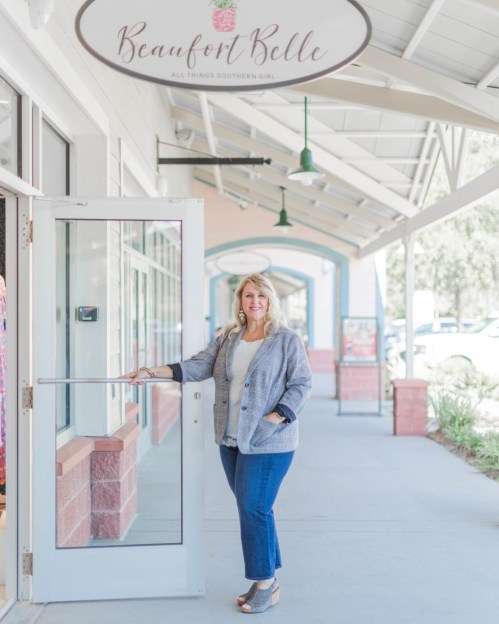A woman stands at the entrance of a boutique, smiling and wearing a stylish outfit, with a sign above reading "Beaufort Belle."