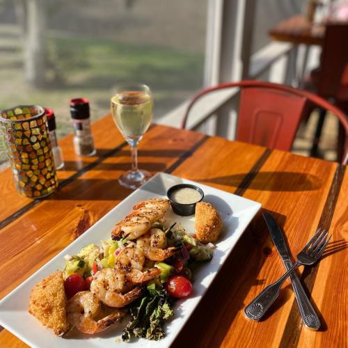 A plate of grilled shrimp with salad and two fried balls, accompanied by a glass of white wine on a wooden table.