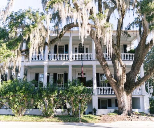 A historic white house with multiple balconies, surrounded by lush greenery and draped with Spanish moss.