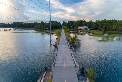 Aerial view of a long, narrow path over water, with trees and power lines in the background, and two people walking.