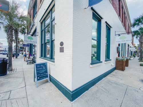 A corner view of a white brick building with green accents, featuring signs for "Tidal Creek Haven" and a chalkboard menu.