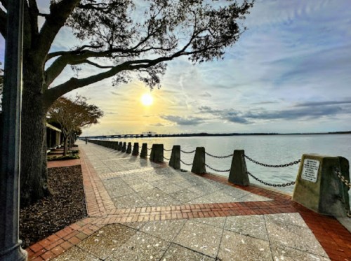 Scenic waterfront path at sunset, lined with trees and a chain barrier, reflecting calm waters and a cloudy sky.