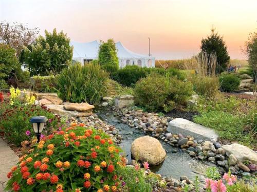 A serene garden with a stream, colorful flowers, and a white tent in the background under a sunset sky.